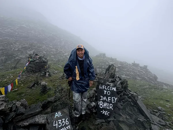 Trekker in a rain cape beside stone cairns and a sign reading Dafey Bhir, 4,336 m